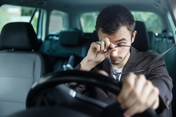 Sleepy young man rubs his eyes with his right hand. His left hand is on the steering wheel. He is sitting at his car.