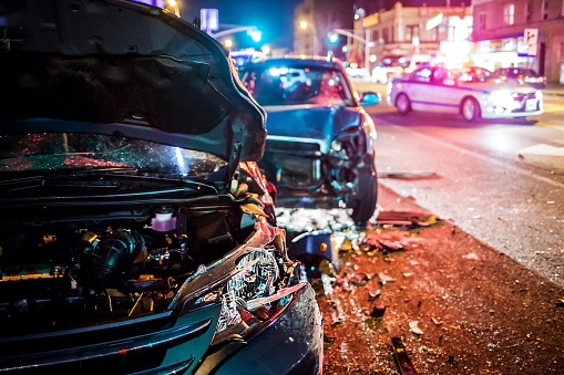 A crash scene at night in the city. Two smashed vehicles on the side of the road with a police car blocking traffic in the street.